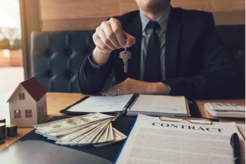 An agent handing house keys across a desk with a lump of money