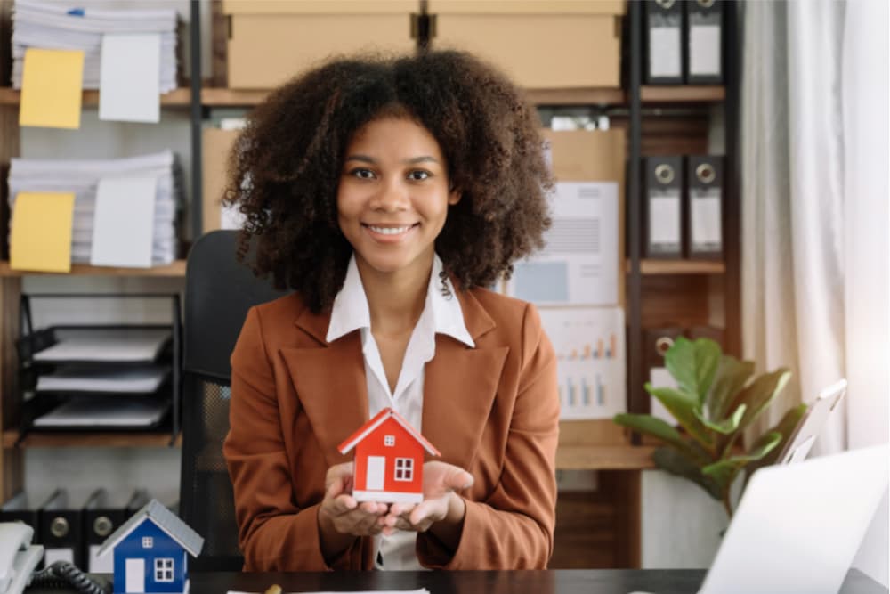 An image of a woman sat at a desk holding a red wooden house model
