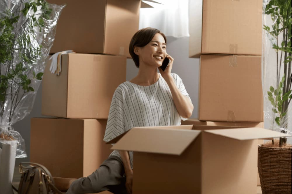 A woman on the phone smiling surrounded by moving boxes stack very high