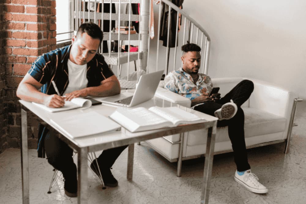 An image of two people sat working on a phone and laptop on a couch and desk