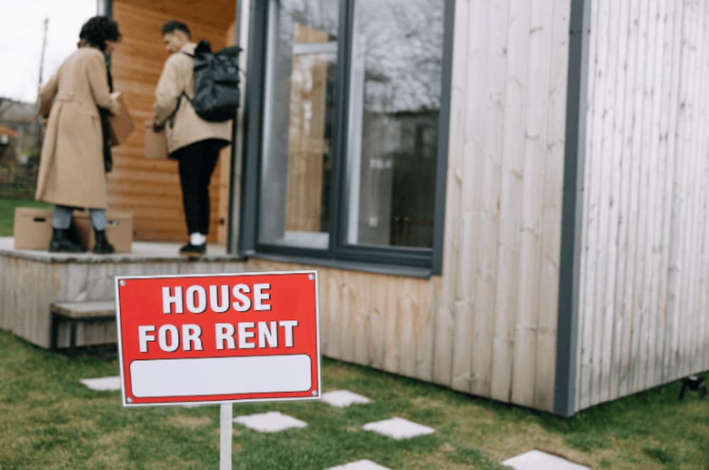 An image of a house for rent sign in the foregound of a house with a couple walking inside