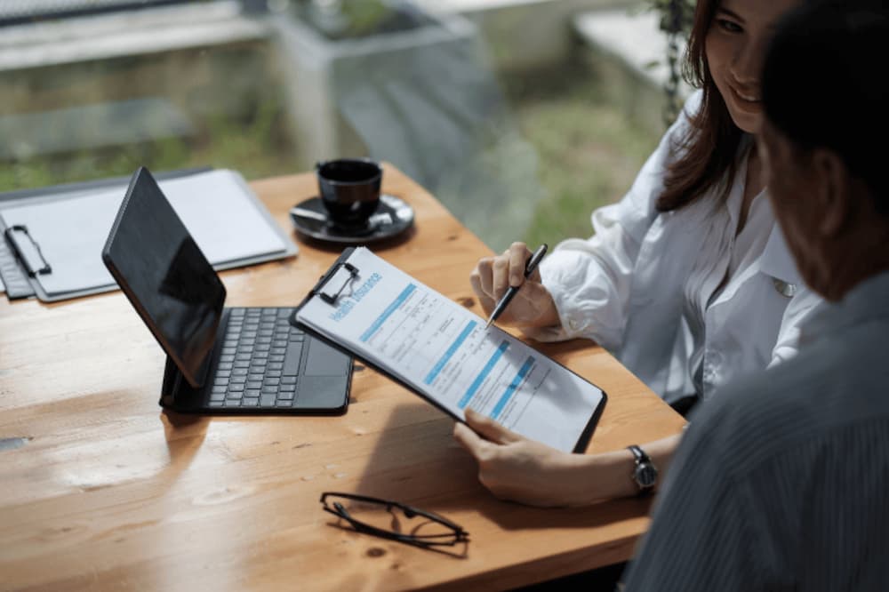 An image of two people discussing health insurance at a desk with a tablet device, clipboard and documents
