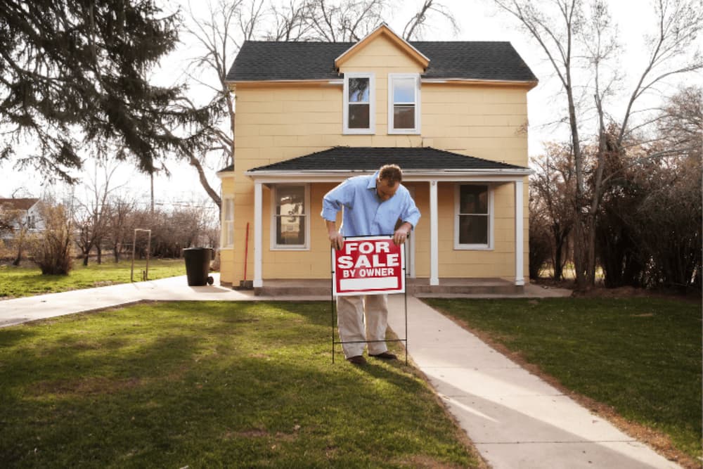 An image of a house with a man putting a for sale sign infront of it