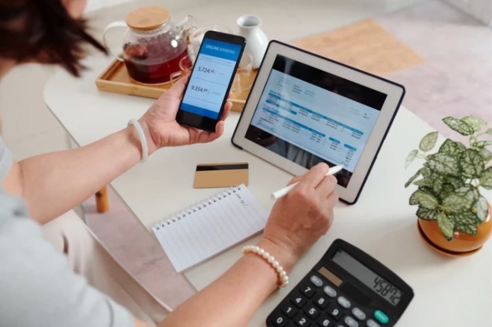 A person sat at a desk checking finance expenses on a phone and tablet with a calculator and pen