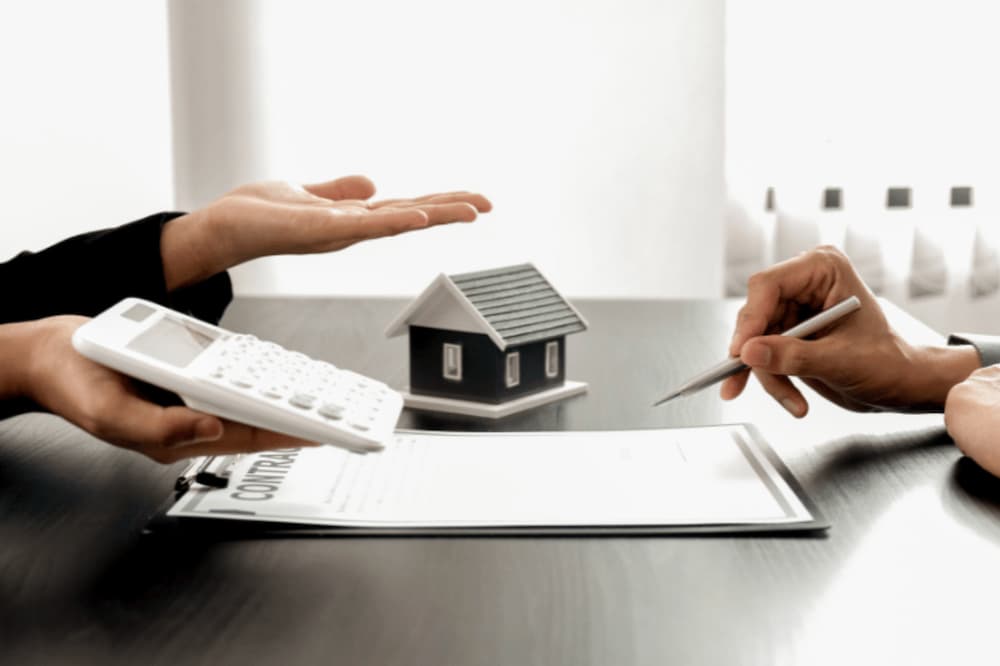 An image of two people sat across a desk one with their hand open with a calculator the other olding a pen above a document
