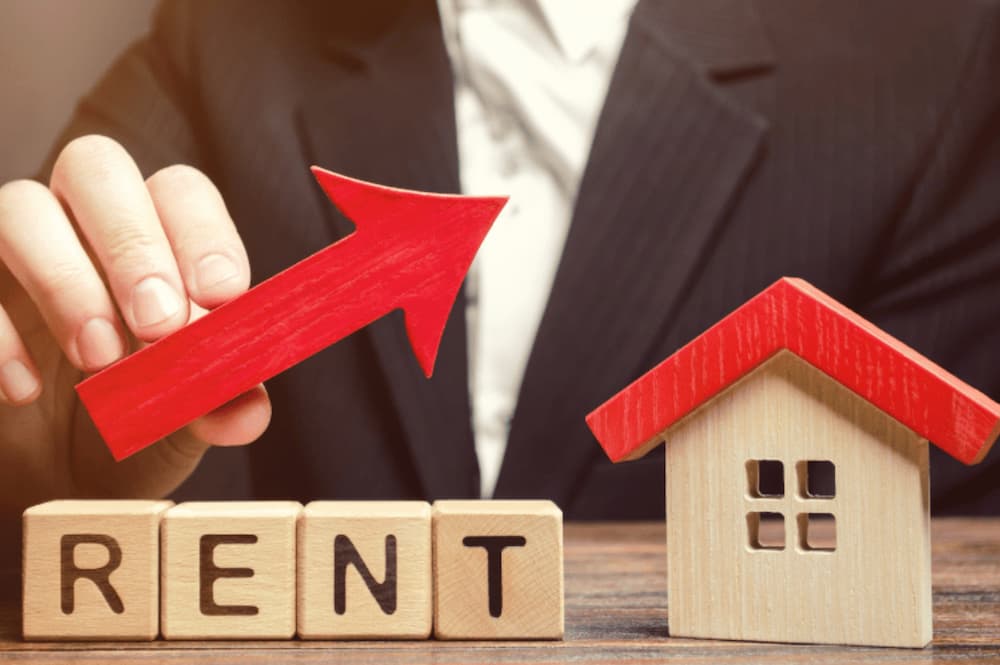 An image someone holding up a wooden arrow pointing upwards above wooden blocks spelling out rent next to a wooden house model